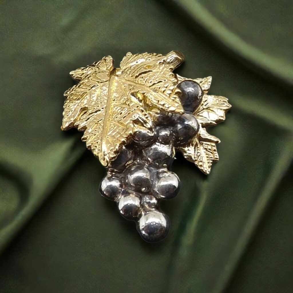 broche en forme de grappe de raisin argentée surmontée de feuilles de vigne dorées, posé sur un tissu vert.